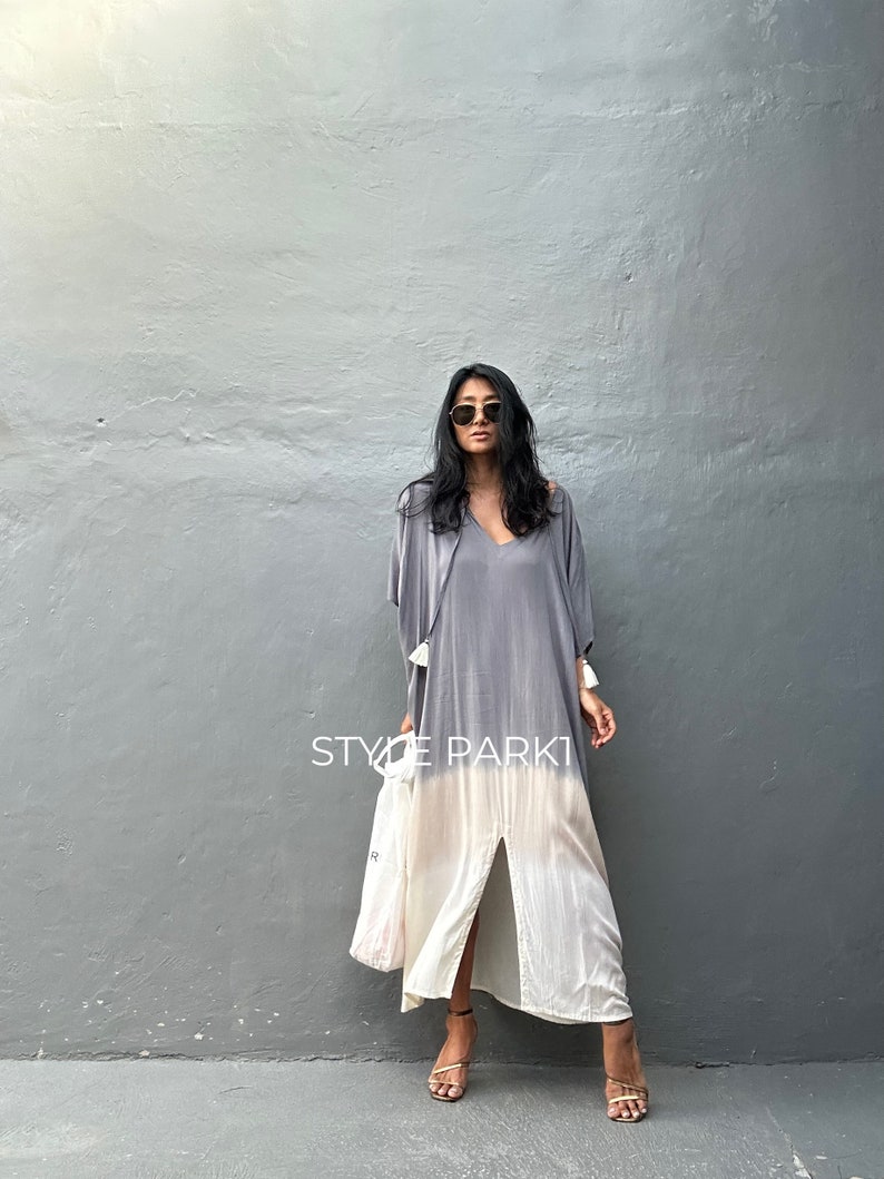 May include: A woman wearing a gray and white ombre maxi dress with a front slit and a tassel detail on the sleeve. The dress is styled with a white tote bag and gold sandals. The text "STYLE PARK1" is visible on the image.