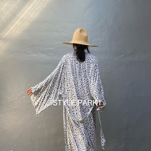 May include: A white and black leopard print kimono with a wide, flowing silhouette. The kimono has long sleeves and a tie belt. The model is wearing a wide-brimmed straw hat.