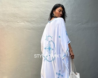 May include: White embroidered kimono with blue floral designs. The kimono has a long, flowing silhouette and is perfect for layering over a simple outfit. The model is wearing the kimono with blue jeans and a white tote bag that says "Style Park".