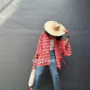 May include: A red and white patterned kimono jacket with wide sleeves. The jacket is worn over a white top and blue jeans. A straw hat and sandals complete the outfit. The text "STYLE PARK" is visible on the jacket.