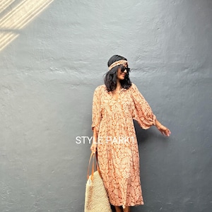 Puede incluir: Un vestido largo y fluido con un estampado floral en tonos melocotón y crema. El vestido tiene mangas largas y cuello en V. Un bolso tote tejido con correas de cuero y sandalias completan el look. El texto "STYLE PARKI" es visible.