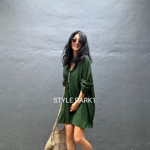 May include: A woman in a dark green dress with long sleeves and a button-down front, carrying a large, beige crocheted tote bag. The text "STYLE PARK1" is visible on the dress. She wears sunglasses and black sandals. The dress is knee-length.