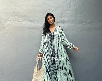 Op de afbeelding: Een vrouw draagt een lange, vloeiende groene en zwarte tie-dye jurk met een V-hals en lange mouwen. Ze houdt een grote, geweven draagtas vast en staat voor een grijze muur. De tekst "STYLE PARK" is zichtbaar op de jurk.