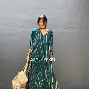 May include: A teal blue and gold tie-dye maxi dress with a V-neck and long sleeves. The dress has a flowing, asymmetrical hemline and is paired with a woven straw tote bag. The text "STYLE PARK1" is visible on the dress.