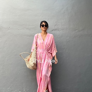 May include: A pink and yellow tie-dye maxi dress with a V-neckline and a side slit. The dress has a tie at the waist and is worn with a straw tote bag and sandals. "STYLE PARK1" is written on the dress.