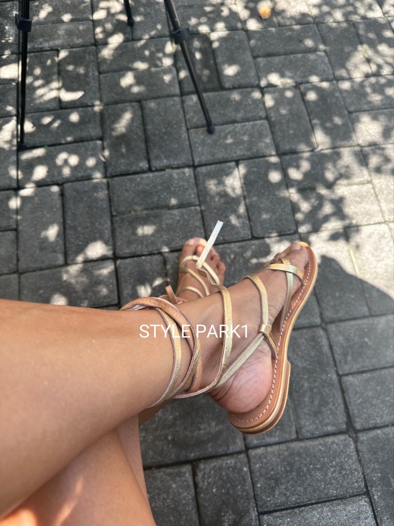 May include: Gold and brown leather sandals with laces that wrap around the ankle. The sandals have a flat sole and are worn on a person's feet. The text "STYLE PARK1" is visible on the sandals.