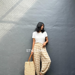 May include: A woman wearing a white t-shirt and patterned pants with a beige tote bag. The pants have a repeating leaf-like design in shades of brown and beige. The tote bag is made of woven straw with brown leather handles. The text "STYLE PARK" is visible.