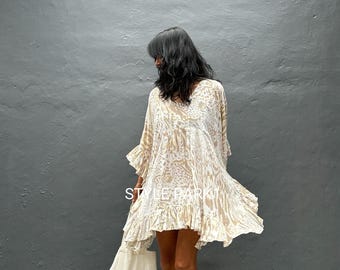 May include: A beige and white patterned dress with a ruffled hem and sleeves. The dress has a V-neck and is knee-length. The text "STYLE PARK" is visible on the dress. The person is wearing gold sandals and carrying a white tote bag.