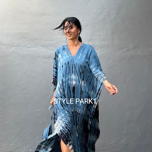 May include: A woman wearing a blue and black tie-dye kaftan with a V-neckline. The kaftan has a long, flowing silhouette and a side slit. The woman is standing in front of a gray wall. The text "STYLE PARK1" is visible on the kaftan.