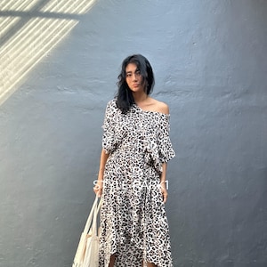 May include: A two-piece outfit featuring a leopard print pattern. The top is off-the-shoulder with short sleeves, and the skirt has an asymmetrical hemline. A canvas tote bag is held in the left hand. The outfit is worn with sandals.