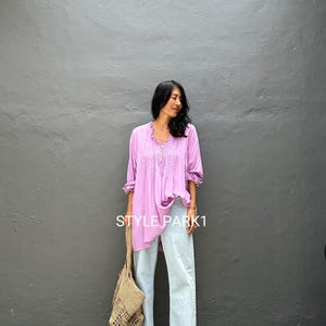 May include: A woman wearing a lilac-colored blouse with a detailed neckline, paired with light-wash denim jeans. She carries a beige woven tote bag. The text "STYLE PARK1" is visible on the blouse. The background is a gray wall.