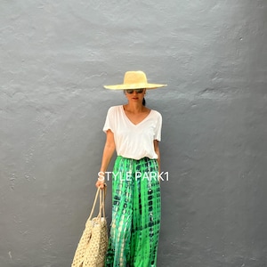 May include: A woman wearing a white V-neck shirt, green tie-dye pants, a straw hat, and sunglasses, holding a woven tote bag. The pants have a vibrant green and black pattern. The text "STYLE PARK1" is visible.