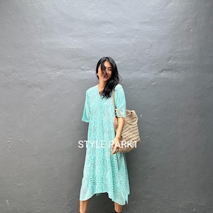 May include: A turquoise midi dress with a white spotted pattern. The dress has short sleeves and a V-neck. The person is wearing black sandals and carrying a woven shoulder bag. The text "STYLE PARK" is visible on the dress.