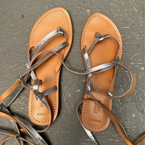 May include: A pair of brown leather sandals with silver straps and laces. The sandals have a simple design and are perfect for summer.