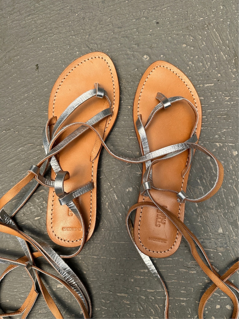 May include: A pair of brown leather sandals with silver straps and laces. The sandals have a simple design and are perfect for summer.