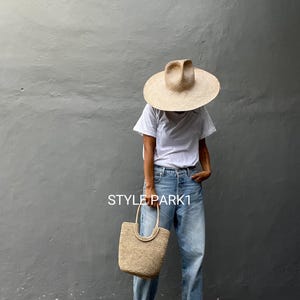 Pu&ograve; includere: Una donna indossa una maglietta bianca, jeans blu chiaro e un cappello di paglia. Porta una borsa a mano in paglia intrecciata. Il testo "STYLE PARK1" &egrave; visibile nell'immagine.