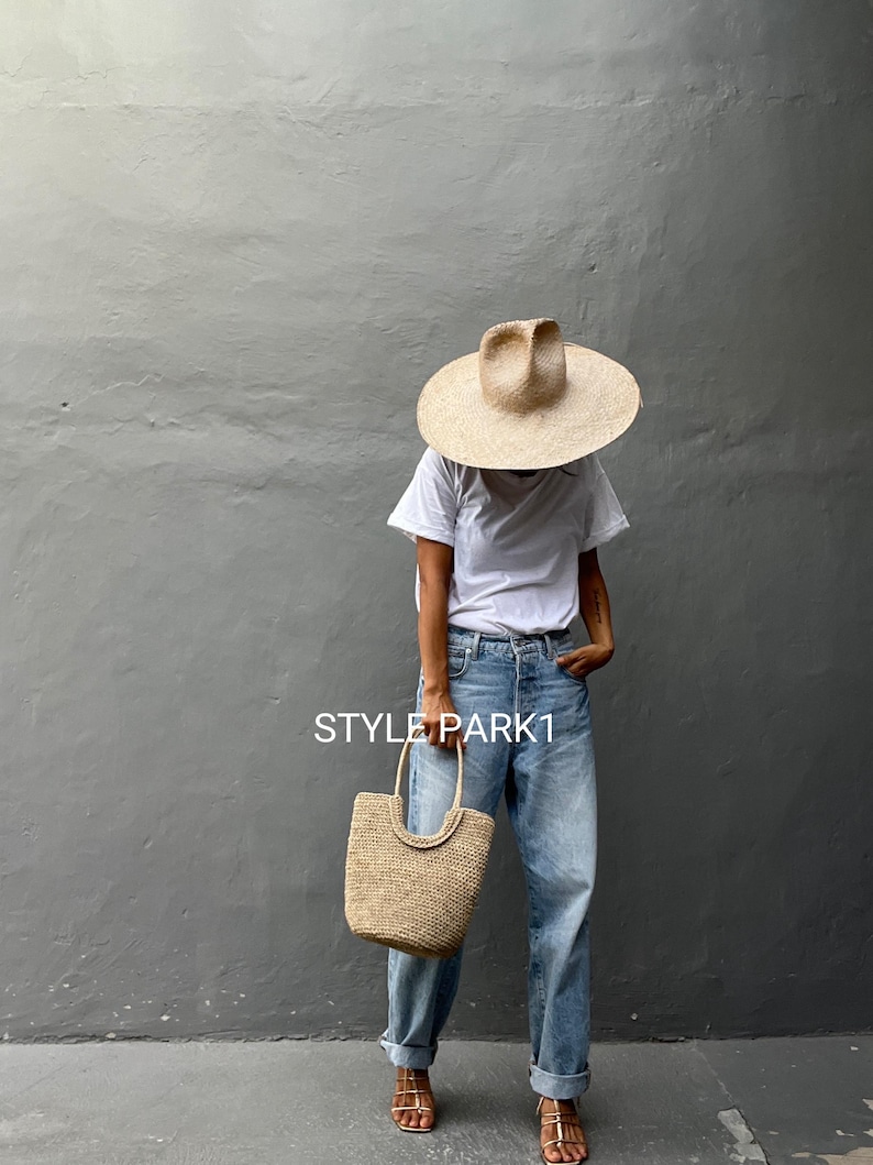 Pu&ograve; includere: Una donna indossa una maglietta bianca, jeans blu chiaro e un cappello di paglia. Porta una borsa a mano in paglia intrecciata. Il testo "STYLE PARK1" &egrave; visibile nell'immagine.
