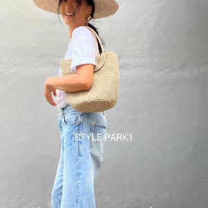 Pu&ograve; includere: Una donna con un cappello di paglia a tesa larga e una maglietta bianca porta una borsa tote in tessuto beige. La borsa ha una tracolla corta e viene indossata sulla spalla. La donna indossa jeans blu e il testo "STYLE PARK1" &egrave; visibile nella parte inferiore dell'immagine.