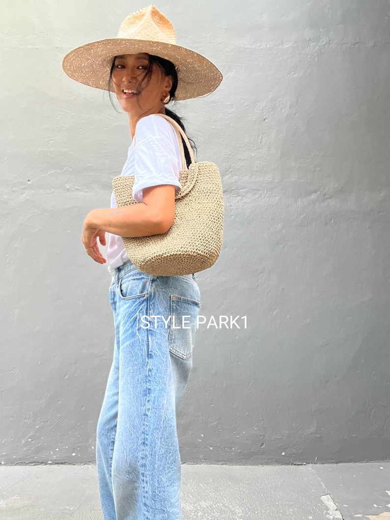Pu&ograve; includere: Una donna con un cappello di paglia a tesa larga e una maglietta bianca porta una borsa tote in tessuto beige. La borsa ha una tracolla corta e viene indossata sulla spalla. La donna indossa jeans blu e il testo "STYLE PARK1" &egrave; visibile nella parte inferiore dell'immagine.