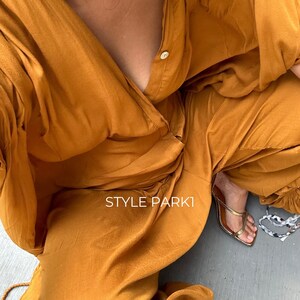 May include: A woman wearing a mustard yellow jumpsuit with a deep V-neckline and a single button closure. The jumpsuit has a relaxed fit and is made of a soft, flowing fabric. The woman is wearing gold sandals and the text "STYLE PARKI" is visible on the jumpsuit.