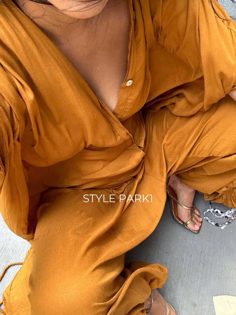 May include: A woman wearing a mustard yellow jumpsuit with a deep V-neckline and a single button closure. The jumpsuit has a relaxed fit and is made of a soft, flowing fabric. The woman is wearing gold sandals and the text "STYLE PARKI" is visible on the jumpsuit.