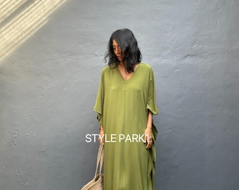 May include: An olive green, flowing dress with a V-neck and short sleeves. The dress is worn with a woven tote bag. The text "STYLE PARK" is visible on the dress. The dress is long and reaches the ankles.