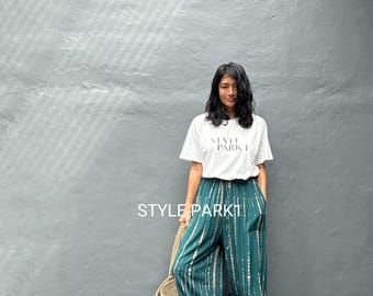 Könnte beinhalten: Eine Frau trägt ein weißes T-Shirt mit der Aufschrift "STYLE PARK I" und eine Hose mit weitem Bein in Türkis und Beige. Sie hält eine gewebte Tragetasche. Der Hintergrund ist eine graue Wand.