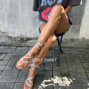 May include: Silver leather sandals with brown straps and laces. The sandals are tied up the legs and feature a criss-cross design. The sandals are worn by a person sitting on a chair. The background is a grey brick wall.