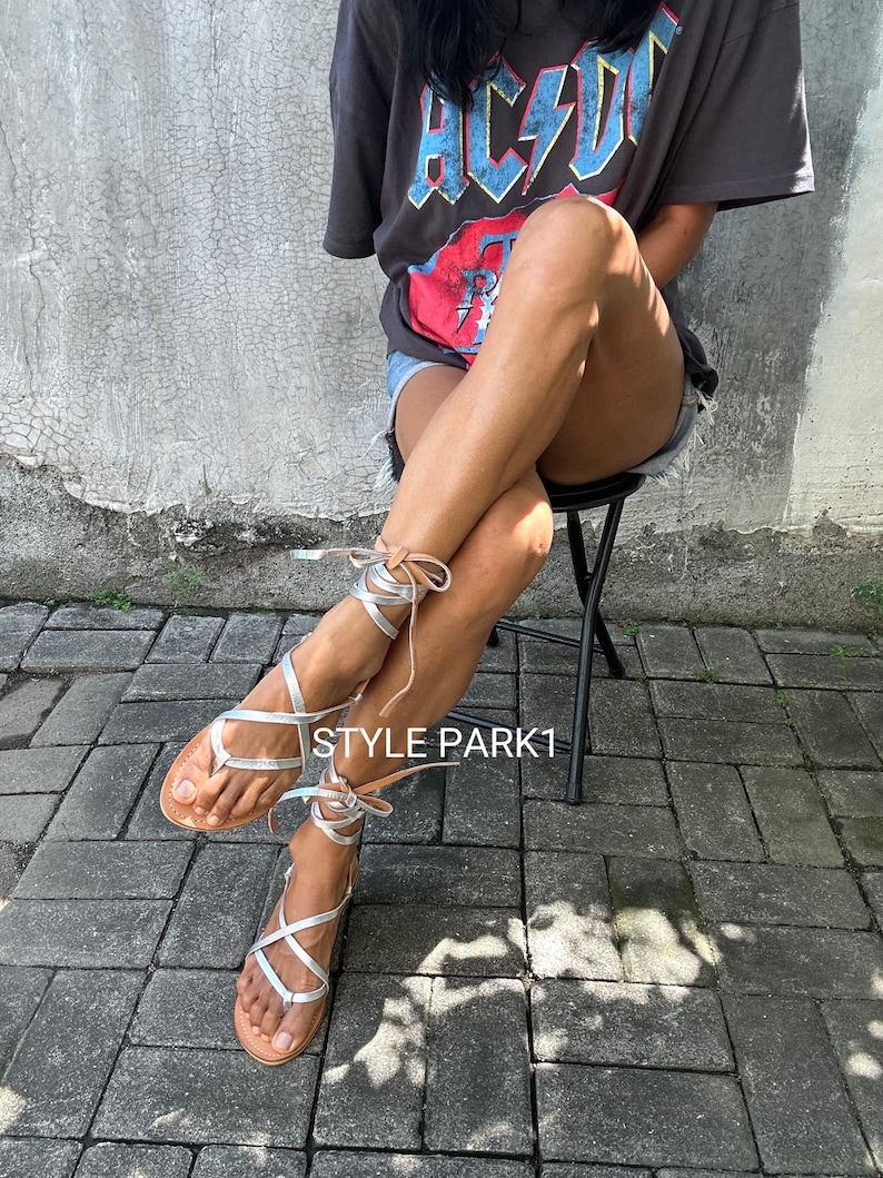 May include: Silver leather sandals with brown straps and laces. The sandals are tied up the legs and feature a criss-cross design. The sandals are worn by a person sitting on a chair. The background is a grey brick wall.