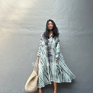 Peut inclure: Une femme portant une longue robe fluide en tie-dye bleu clair et noir avec un col et des manches longues. Elle porte un grand sac fourre-tout en osier et des sandales à talons marrons.