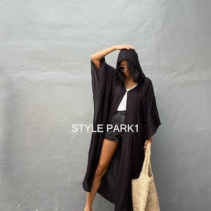 May include: A black hooded long-sleeved dress with a white button closure. The dress is worn with black shorts and gold sandals. The person is carrying a large straw tote bag. The text "STYLE PARK1" is visible on the image.