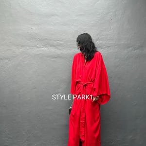 Op de afbeelding: Een lange, vloeiende rode robe met wijde mouwen en een bijpassende riem. De robe is gemaakt van een zachte, draperende stof en wordt over een donkere outfit gedragen. De tekst "STYLE PARK1" is zichtbaar op het kledingstuk.