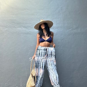 Puede incluir: Una mujer con un top de bikini azul marino, pantalones teñidos con tinte, un sombrero de paja y una bolsa de mano tejida. Los pantalones tienen un estampado blanco, negro y naranja. El sombrero tiene un ala ancha.