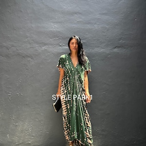 Puede incluir: Un vestido maxi verde con estampado tie-dye, cuello en V y mangas cortas. El vestido tiene una silueta fluida y un patrón marrón y blanco. El texto "STYLE PARK 1" es visible en el vestido. La persona lleva sandalias doradas y sostiene un clutch negro.