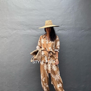 Puede incluir: Una camisa de botones y pantalones entallados a juego en color tostado con un estampado de palmeras blancas, un sombrero de paja de ala ancha, un bolso de paja trenzada y sandalias de tiras. El texto STYLE PARK 1 es visible en blanco.