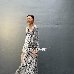 May include: A black and white maxi dress with a large circular pattern and floral print. The dress has long sleeves and a V-neckline. The model is wearing the dress with a straw bag and sandals. The text "STYLE PARK1" is visible on the dress.