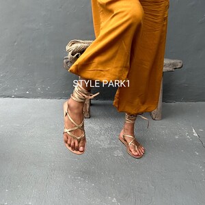 May include: Gold leather sandals with laces that wrap around the ankles. The sandals are worn by a person wearing a mustard yellow wide-leg pant. The text "STYLE PARK1" is visible on the pant leg.