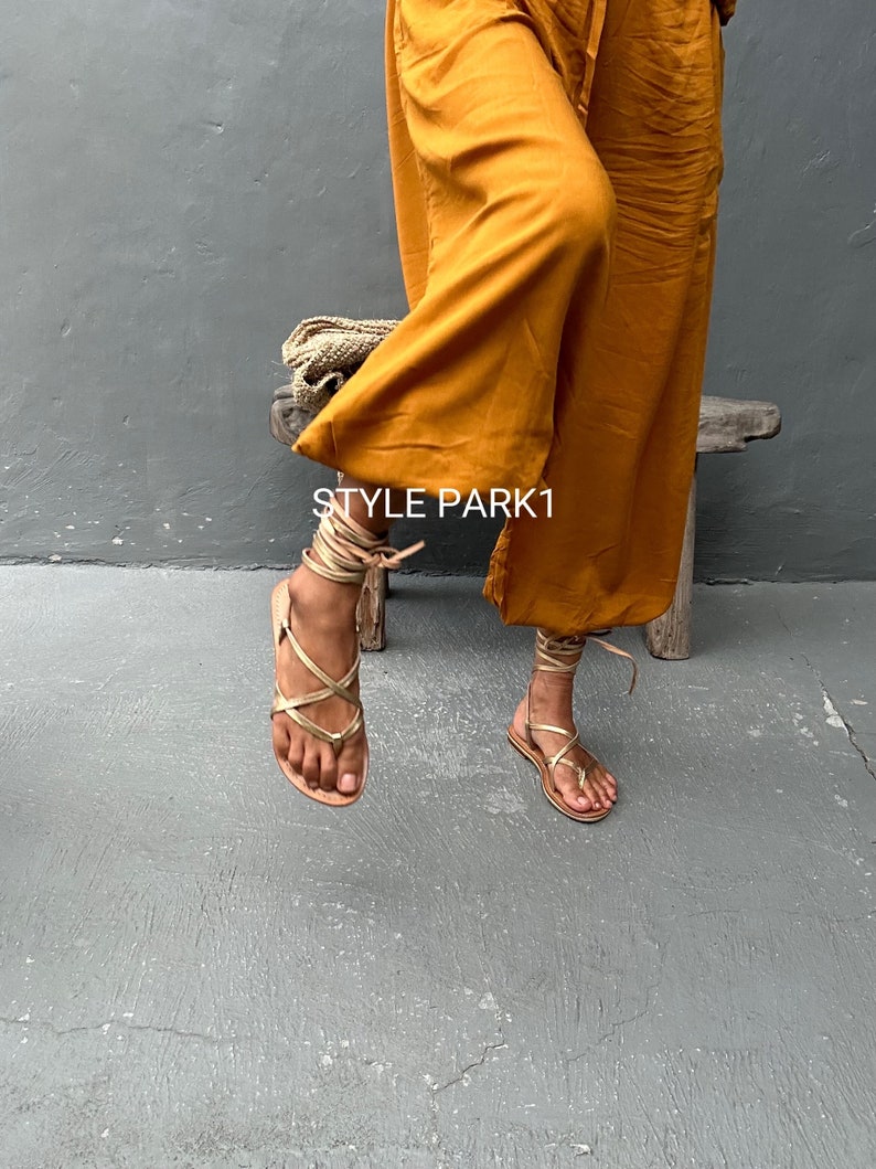 May include: Gold leather sandals with laces that wrap around the ankles. The sandals are worn by a person wearing a mustard yellow wide-leg pant. The text "STYLE PARK1" is visible on the pant leg.