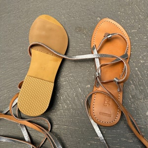 May include: A pair of brown leather sandals with silver straps and laces. The sandals have a non-slip sole and the brand name "STYLE PARIS" is printed on the sole.