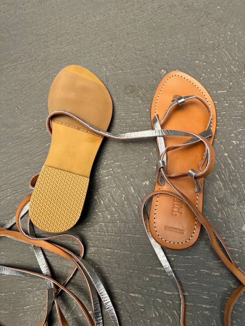 May include: A pair of brown leather sandals with silver straps and laces. The sandals have a non-slip sole and the brand name "STYLE PARIS" is printed on the sole.