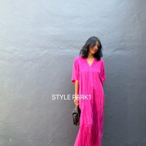 Pode incluir: Uma mulher vestindo um vestido longo rosa com decote em V, uma bolsa preta e sandálias prateadas. O vestido tem um visual texturizado e enrugado. O texto "STYLE PARK1" é visível na imagem.