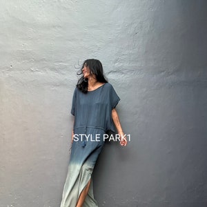 Puede incluir: Un vestido largo y fluido, de color degradado, con la parte superior azul oscuro y la inferior azul claro. El vestido tiene un lazo en la cintura y una abertura lateral. La modelo lleva el vestido con sandalias y está de pie frente a una pared gris. El texto "STYLE PARK1" es visible en el vestido.