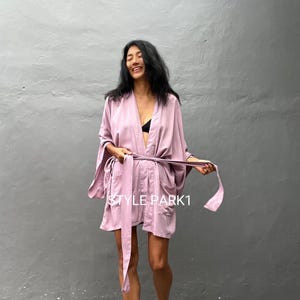 Op de afbeelding: Een mauvekleurige kimono-jas met een strikband. De jas is gemaakt van een zachte, zijdeachtige stof en heeft een ontspannen pasvorm. Het model draagt de jas met een zwarte bh eronder.