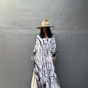 May include: A white and black tie-dye maxi dress with a V-neckline and long sleeves. The dress is worn with a wide-brimmed straw hat and gold sandals. The text "STYLEPARK1" is visible on the dress.