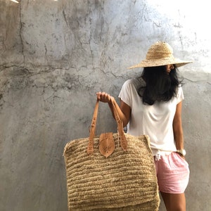 May include: A large, woven straw tote bag with leather handles and a decorative leather flap. The bag is tan and beige, with a rectangular shape. The person is wearing a straw hat, white shirt, and red and white shorts.