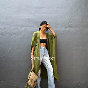 May include: An olive green open-front kimono with wide sleeves, worn over a black bandeau top and light-wash blue denim jeans. The outfit includes a woven tan straw handbag with a long strap. The text STYLE PARK1 is visible on the image.