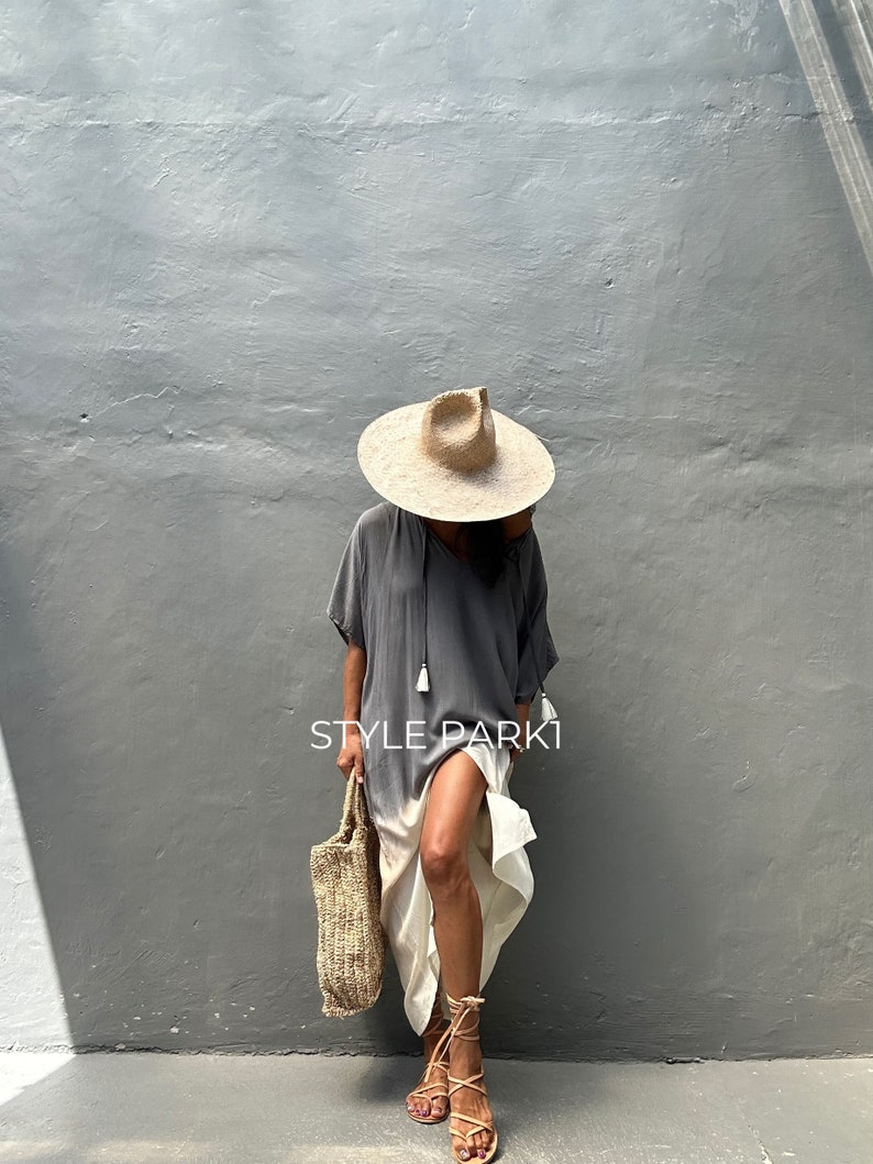 May include: A woman wearing a gray and white ombre maxi dress with a straw hat and sandals. She is carrying a woven tote bag. The text "STYLE PARK1" is visible on the dress.