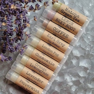 May include: A selection of lip butters with flavours such as Matcha Green Tea, Lemon Beet Root, and Coffee. Each lip butter has a brown label indicating its flavour. Dried lavender sprigs are in the background, adding a natural touch.