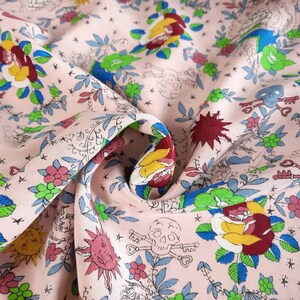 May include: Pink fabric with a repeating pattern of roses, skulls, keys, and suns. The floral and skull design is in shades of red, yellow, blue, and green. The fabric has a slightly wrinkled texture.