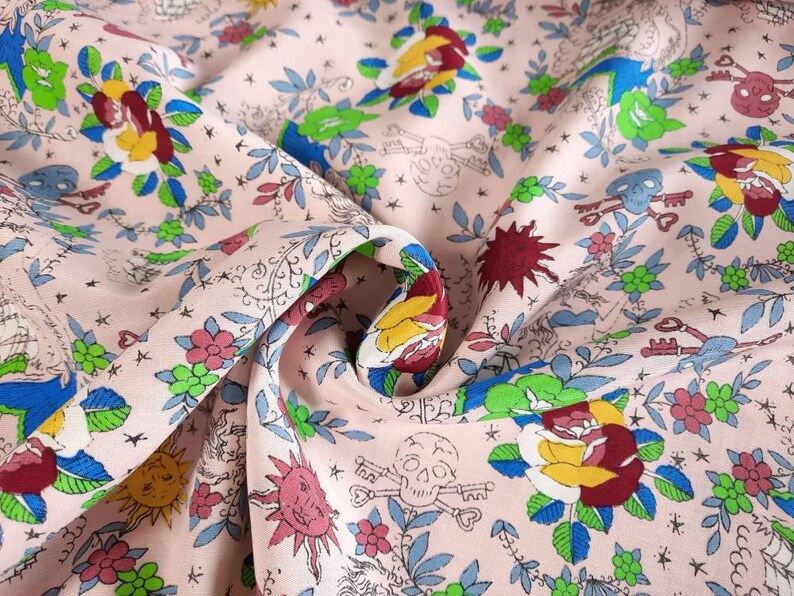 May include: Pink fabric with a repeating pattern of roses, skulls, keys, and suns. The floral and skull design is in shades of red, yellow, blue, and green. The fabric has a slightly wrinkled texture.